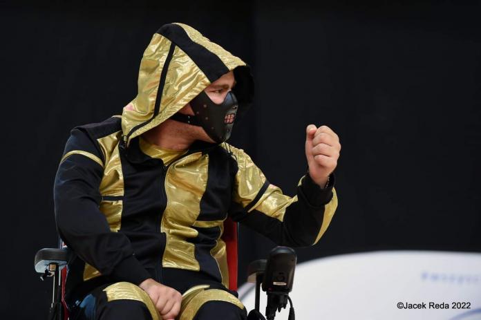 A dancer in a Hannibal Lecter mask and in a black and gold costume pumps his left fist during a dance.