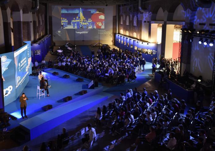 Two people speak from the stage in front of a packed auditorium.