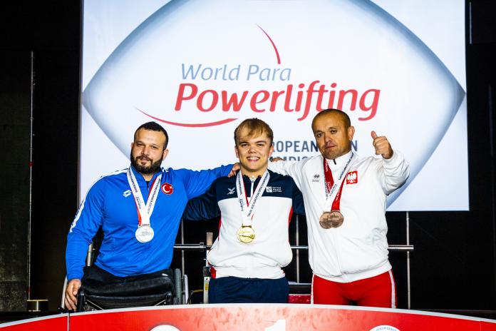 Three male Para powerlifters with double medals around their necks pose together on the podium.