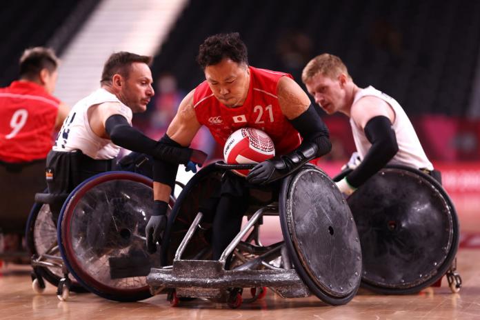 A male wheelchair rugby player in a red jersey carries the ball past two male athletes.