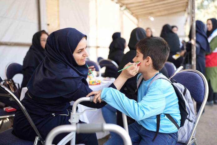 A woman paints the Iranian flag on the left cheek of a young boy.