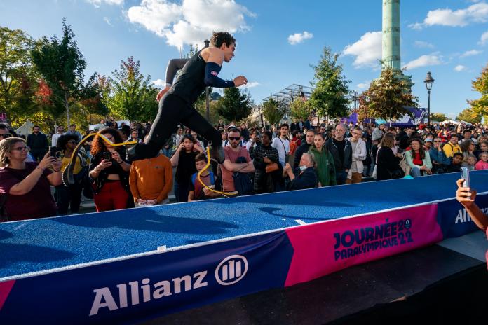 A male long jumper on two blades runs along a blue track towards a sandpit.