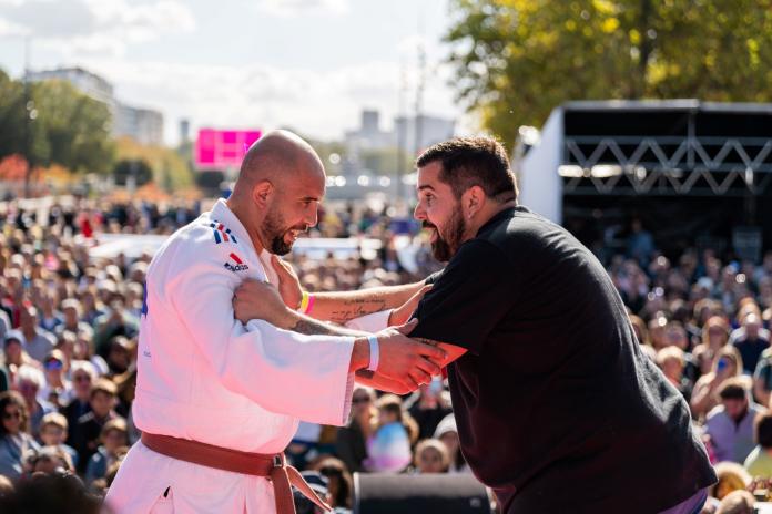 An athlete in judo uniform grapples with a spectator on a stage.