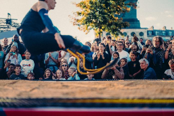 A male long jumper on two blades jumps into a sandpit in front of a large crowd.