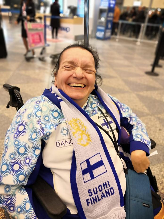 A female laughs as she sits in an electric wheelchair with a Finland scarf around her neck.