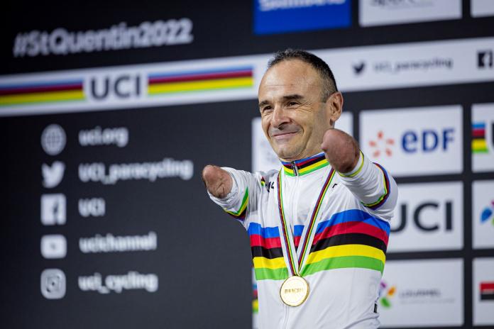A male cyclist who is missing his arms above the elbow celebrates on the podium with a gold medal.