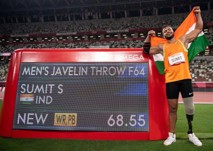 A male athlete holds the Indian flag and stands next to a screen showing his name and record