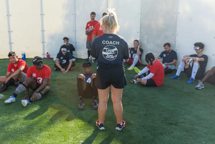 A female coach stands in front of male blind soccer players