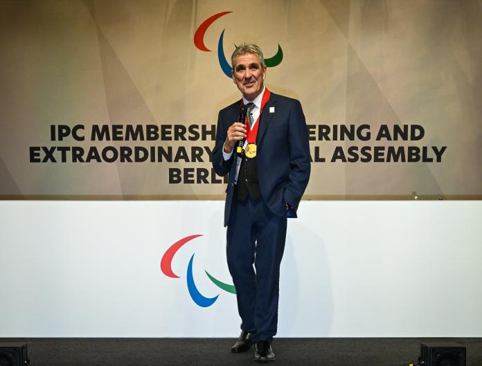 A man with a gold medal around his neck gives a speech from the stage with the Agitos logo behind him.