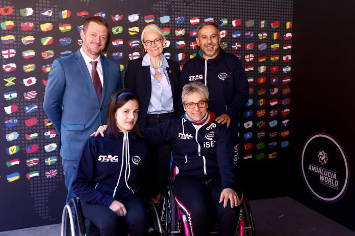 Five people, including two females in wheelchairs, pose by the official background of the Andalucia World Para Table Tennis Championships.