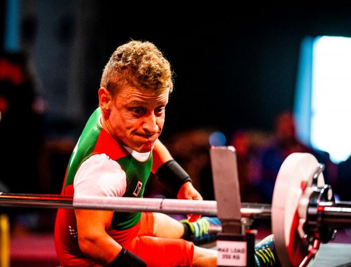 A female athlete of short stature turns back on the bench to look at the barbell with a look of determination on her face.