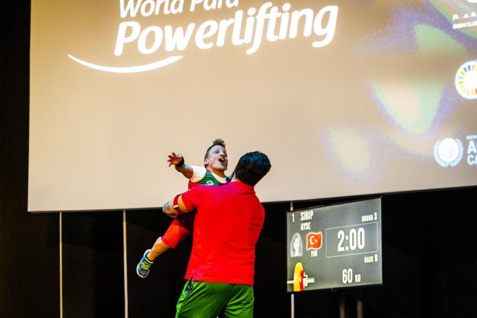 A female athlete of short statue is lifted off the ground by her coach as she celebrates with her arms spread wide.