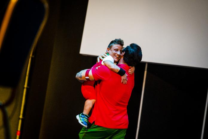 A female athlete of short statue smiles as she is lifted off the ground by her coach for a post-competition hug.