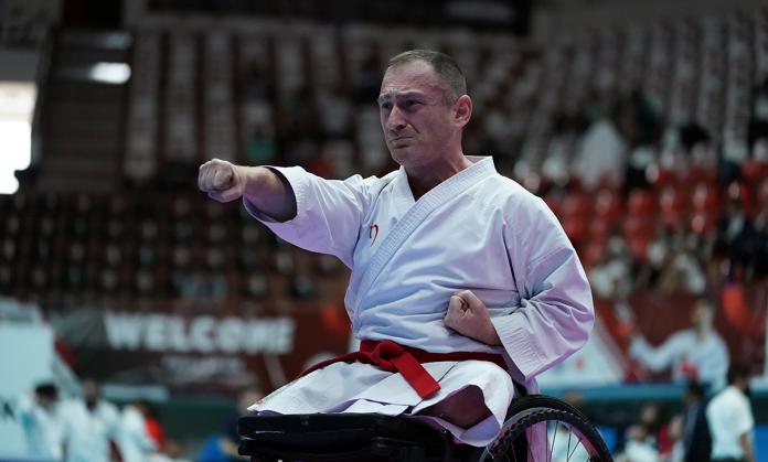 A male athlete on a wheelchair, who is wearing a white karate uniform, punches with his right fist during a Para karate competition