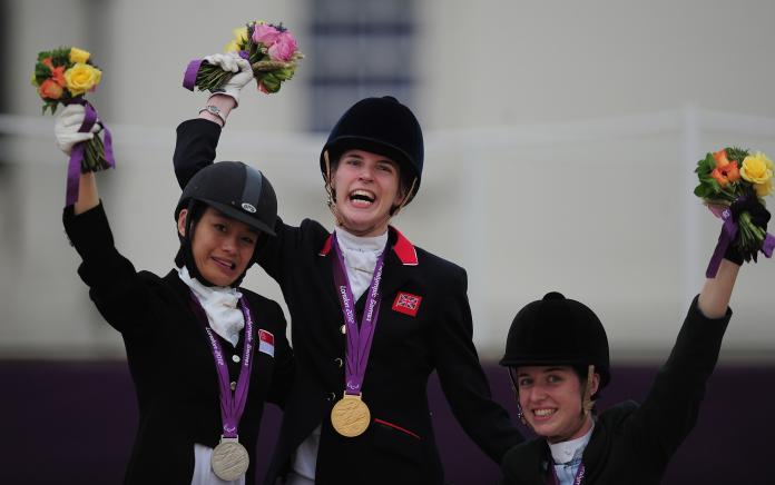 Three female Para equestrian riders with medals around their necks wave bouquets while on the podium.