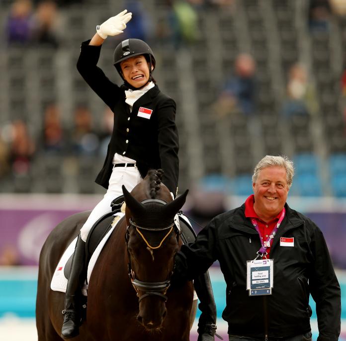 A female Para equestrian rider waves while sitting on a horse as her coach leads the horse away by the reins.