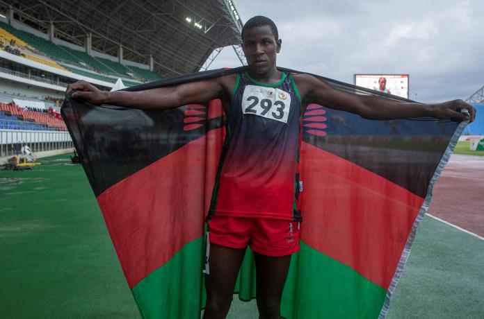 A male athlete spreads out his arms on the track while holding the Malawi flag.