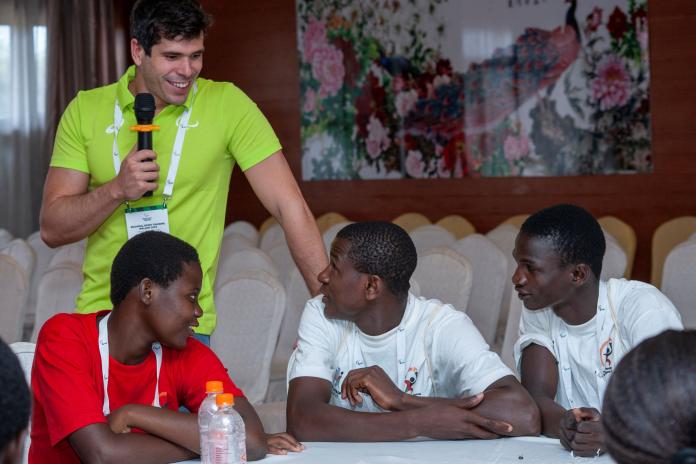 A male athlete in a green shirt with an Agitos logo holds a microphone as he smiles and looks towards a table of three Malawian athletes.