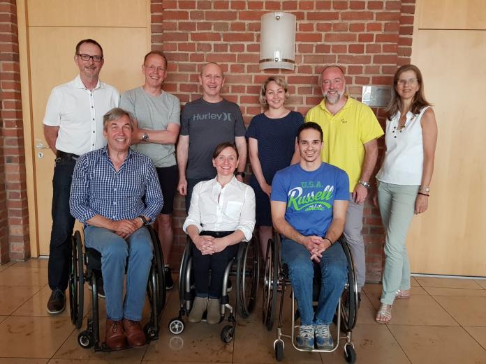 Nine members of the IPC Medical Committee, three in the front row in wheelchairs and six standing in the back row, smile as they pose for a group photo.