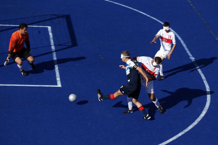 A man in a blindfold shoots at goal in a blind football match at the London 2012 Paralympic Games while a player from the opposing team tries to hold him back.