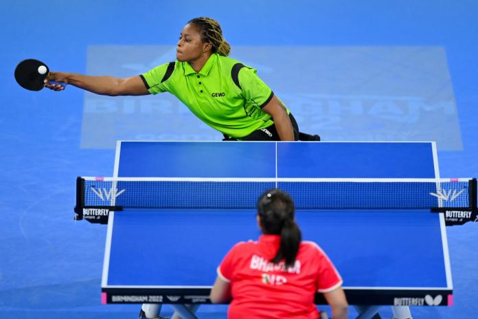 A female table tennis player returns a shot during a match, right arm stretched wide and her wheelchair tilted as her opponent looks on.