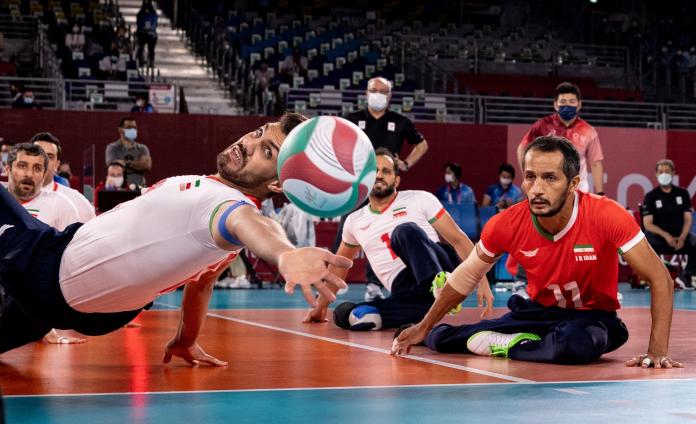 A male sitting volleyball player stretches out his left arm and flips backwards as he attempts to return the ball.