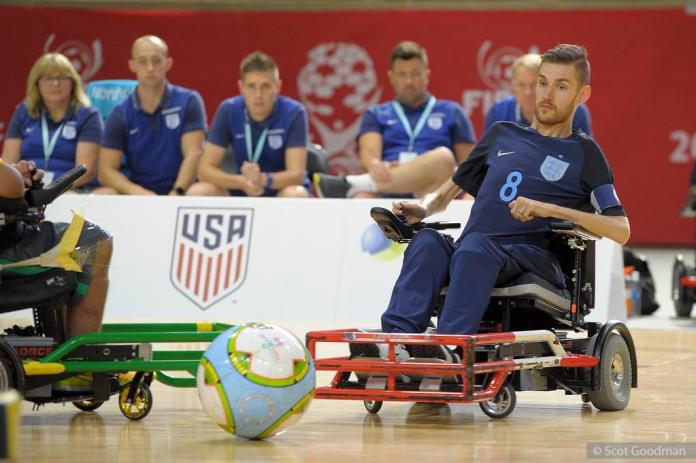 A male powerchair football player controls his electric wheelchair in front of the ball, while four people watch the game from outside of the court.