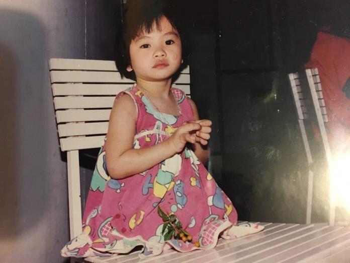 A young girl in a pink dress and without legs sits on a deck chair in a vintage family photo.