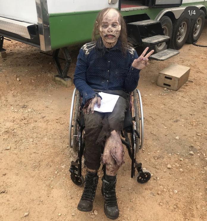 A female actress in full zombie costume and makeup gives a peace sign in an informal photo on a film set.