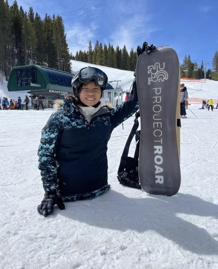 A female athlete without legs below the hip stands on snow while holding up a snowboard with the words 'Project ROAR' written on it.
