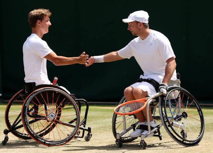 Two male wheelchair tennis players clap hands after getting a point in a match on a grass court.