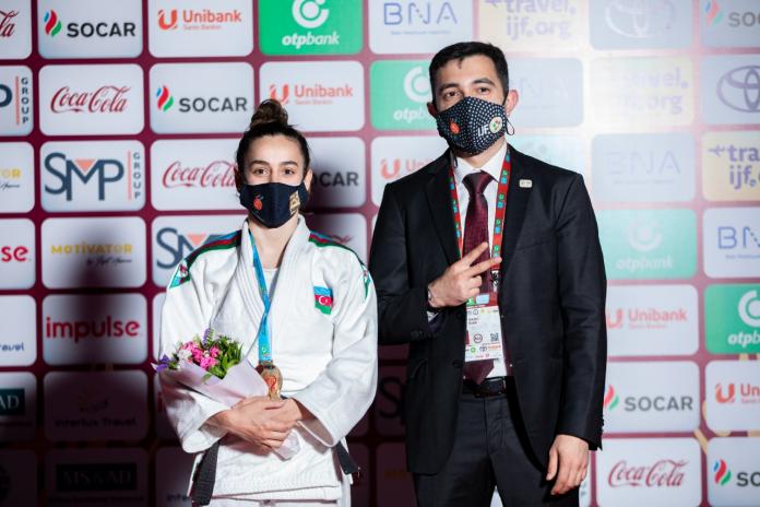 A female judoka in a competition uniform, with medal and flowers, poses by an official backdrop next to a man in a suit.