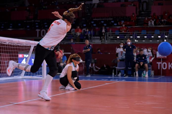 A female goalball player throws the ball while her teammate sits on the competition floor, prepared to block the returning shot.