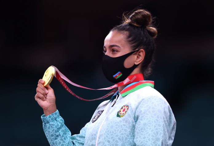 A female athlete in national kit and a mask with an Azeri flag holds up her gold medal as she poses for photos.