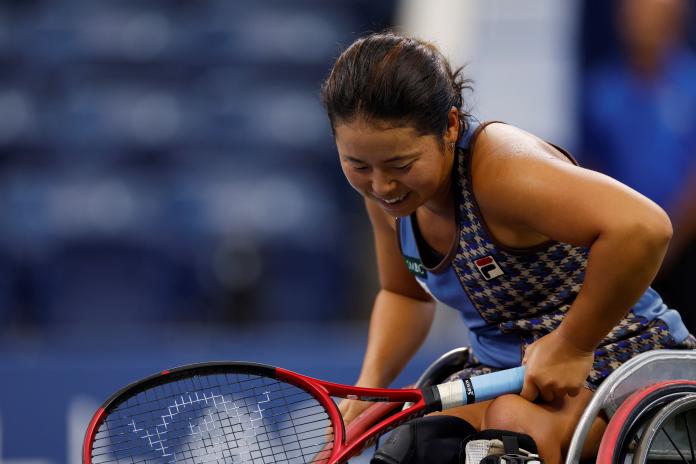 A female wheelchair tennis player looks down with a small smile during a pause in the match.