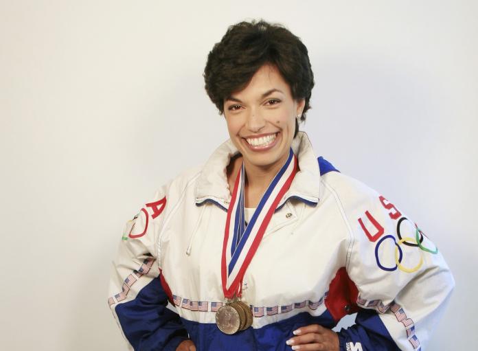 A woman, wearing a white Team USA jacket, poses for a photograph with three medals around her neck
