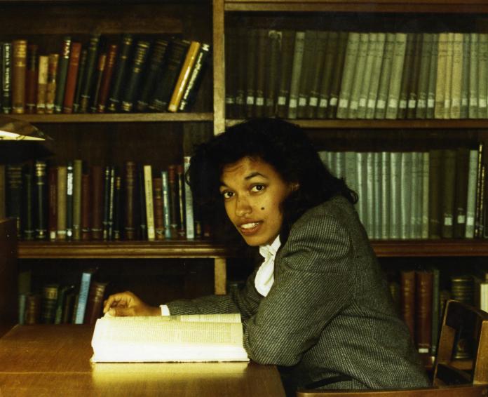 An African-American woman sits in front of a bookshelf and reads