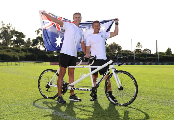 A male athlete and his guide hold the Australian flag and their tandem bike