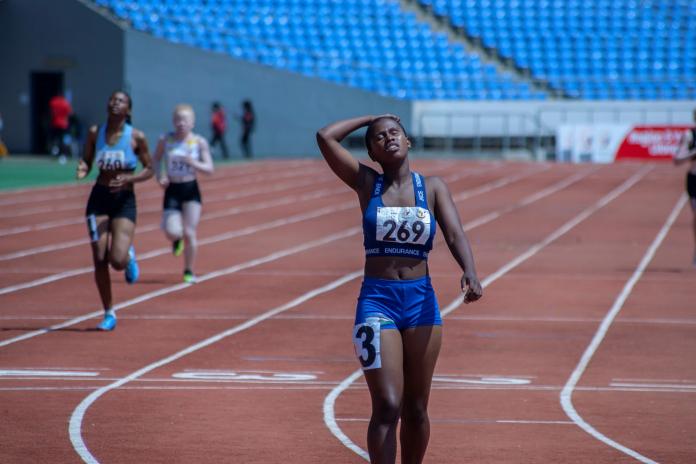 A female sprinter dips her head back and clutches it with her right hand as she recovers from a race finish. 