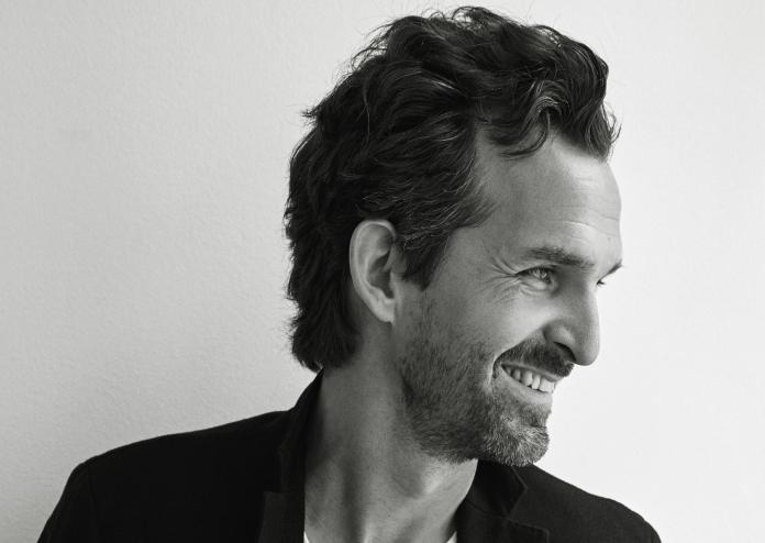 A black and white portrait of a man in his 40s, smiling as he looks away from the camera.