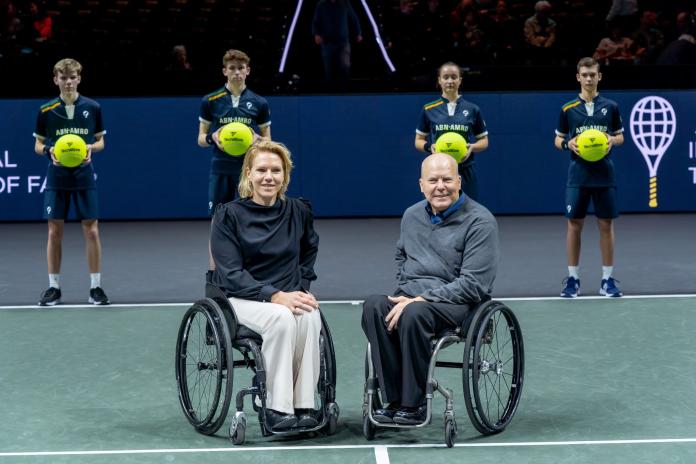 A woman and a man sit in wheelchairs on a hard green court and pose for an official photo.
