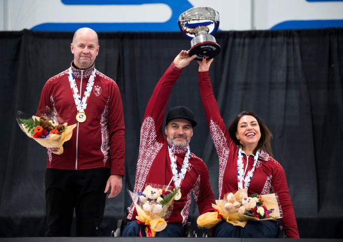 A female and a male athlete lifts a trophy together, while a male coach stands beside them.