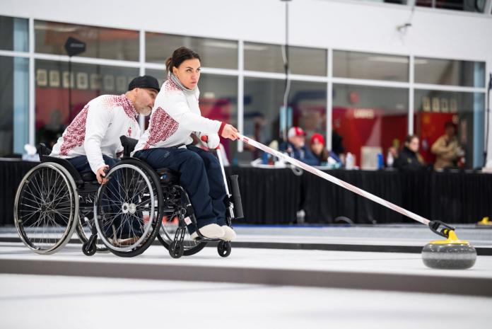 A female wheelchair curler delivers a stone with a stick, while a male athlete holds onto her wheelchair
