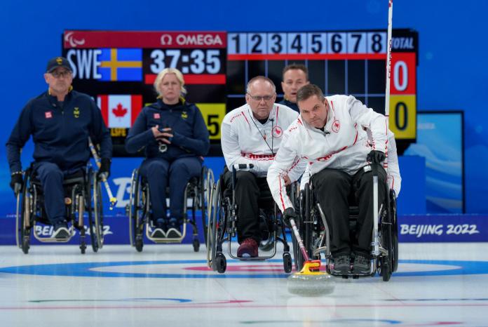 A male wheelchair curling athlete slides a stone, while four athlete watch him on the ice