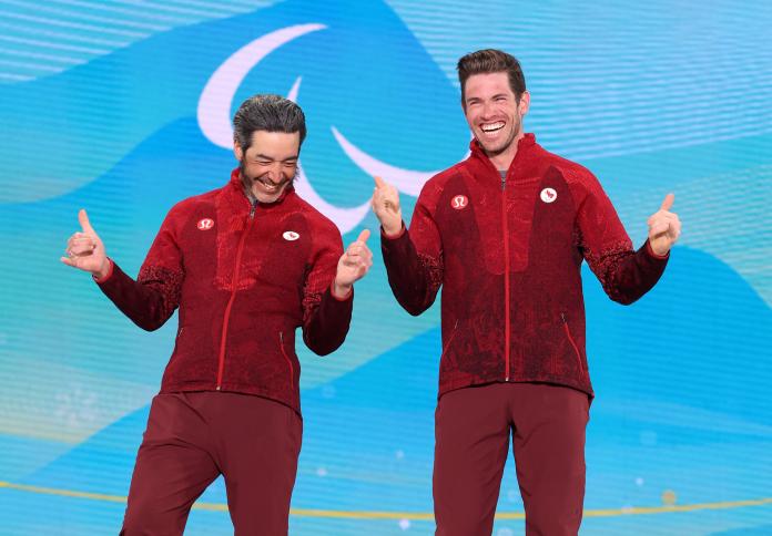 Two athletes in Canadian kit make celebratory gestures with their hands during a medal ceremony.