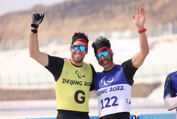 Two men, an athlete and his guide, wave as they celebrate a golden finish in the sprint race.