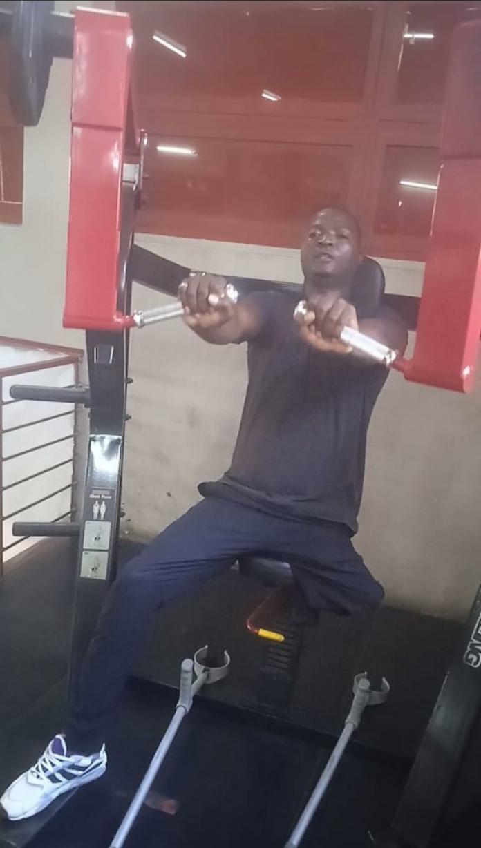 A male Para powerlifter without a left leg above the knee lays down his crutches to press weights on a lifted bench.