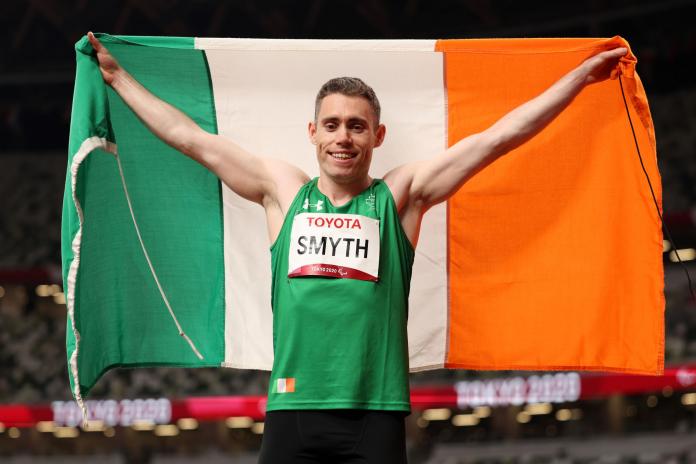 Un atleta masculino sostiene una bandera irlandesa detrás de él mientras celebra una victoria en el estadio de atletismo en Tokio 2020.