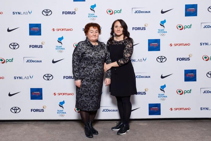 Two women in evening wear hold hands and pose for a photo next to an official backdrop.