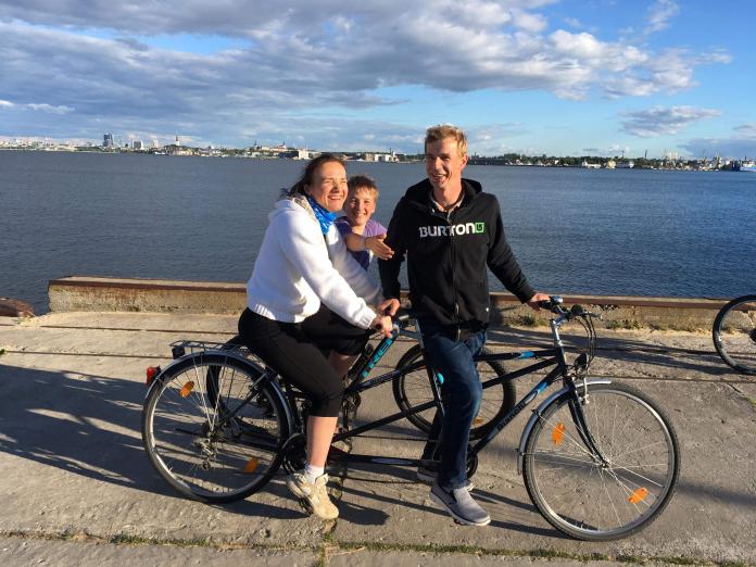 A man and woman pause during a tandem bike ride with a young boy, also on a bike, stopping and smiling behind them.